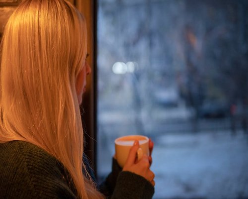 Person thoughtfully looking out the window with a coffee cup
